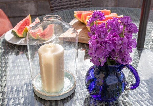 Ein Tisch mit einem Glaskerzenhalter, einer Vase mit violetten Blumen und einem Teller mit Wassermelone.