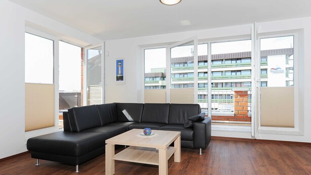Ein modernes Wohnzimmer mit einer schwarzen Eckcouch, einem Holztisch und großen Fenstern mit Blick auf ein Gebäude mit Balkonen.