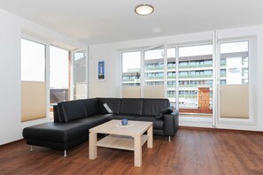 A modern living room with a black sectional sofa, a wooden coffee table, and large windows offering a view of a building with balconies. - Red sand - house lighthouse (Esens)