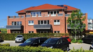 Ein mehrstöckiges Backsteinhaus mit rotem Dach und mehreren Balkonen, sowie ein Parkplatz mit einigen Autos.