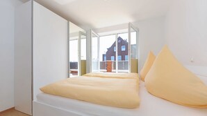 Ein modernes Schlafzimmer mit einem Bett, weißen Wänden und einem großen Fenster mit Blick auf ein Backsteinhaus.