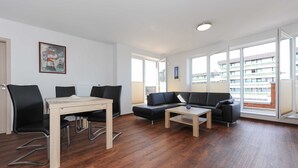 Ein modernes Wohnzimmer mit Esstisch, schwarzen Stühlen, einer schwarzen Couch und einem Couchtisch. An der Wand hängt ein Gemälde und große Fenster sorgen für natürliches Licht.