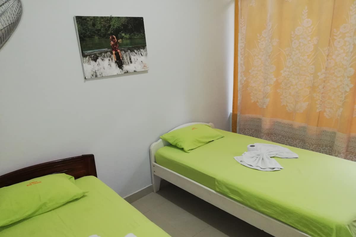 Quarto pequeno e limpo com duas camas, um quadro na parede e uma cortina.