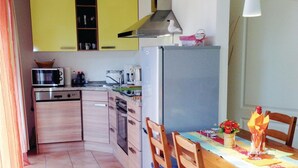 Een kleine keuken met een magnetron, fornuis, spoelbak en een koelkast. Er is een eettafel met stoelen en een vaas met bloemen.