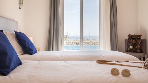 A hotel room with a large bed, a view of the sea, and a chair with a bag.