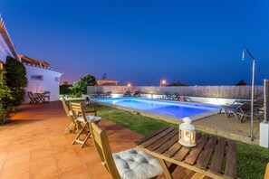 Piscina con sillones de descanso y una mesa, rodeada por un patio con suelo de baldosas y un pequeño jardín.