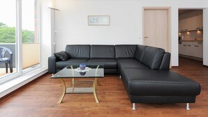A modern living room with a black leather sectional sofa, a glass coffee table, and a view of the kitchen through an open door. - Stader Sand - House Lighthouse (Esens)