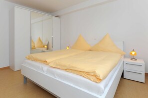 Un lit bien fait, avec un cadre blanc et une literie jaune, une table de chevet blanche avec une lampe, et un grand miroir accroché au mur.