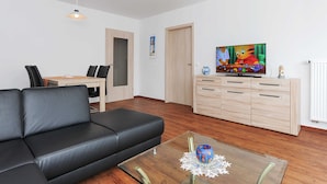A modern living room with a black leather sofa, a glass coffee table with a decorative item, a wooden cabinet with drawers, a television displaying a cartoon, and a dining area with a table and chairs. - Stader Sand - House Lighthouse (Esens)
