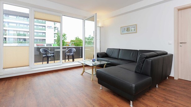 A modern living room with a black leather sofa, a glass coffee table, and a view through large sliding glass doors to an outdoor balcony with a chair. - Stader Sand - House Lighthouse (Esens)
