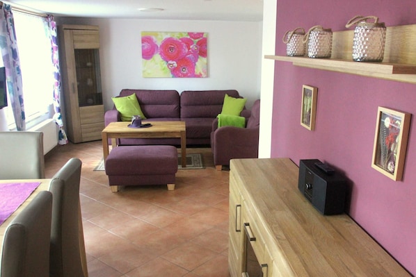 A living room with a sofa set, a coffee table, and a TV. There is a cabinet with a TV on top, a shelf with decorative jars, and a painting on the wall.