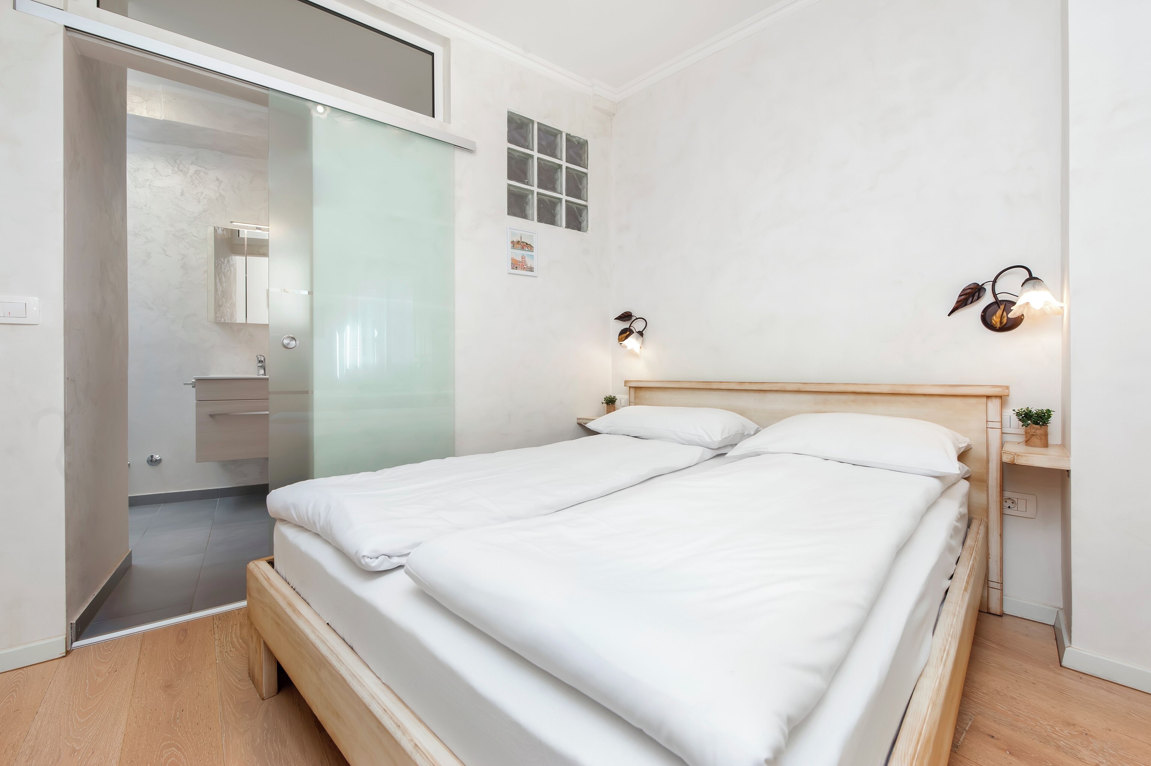 Une chambre à coucher avec un lit en bois, une table de chevet, une lampe fixée au mur et une petite étagère sur laquelle est posée une plante.