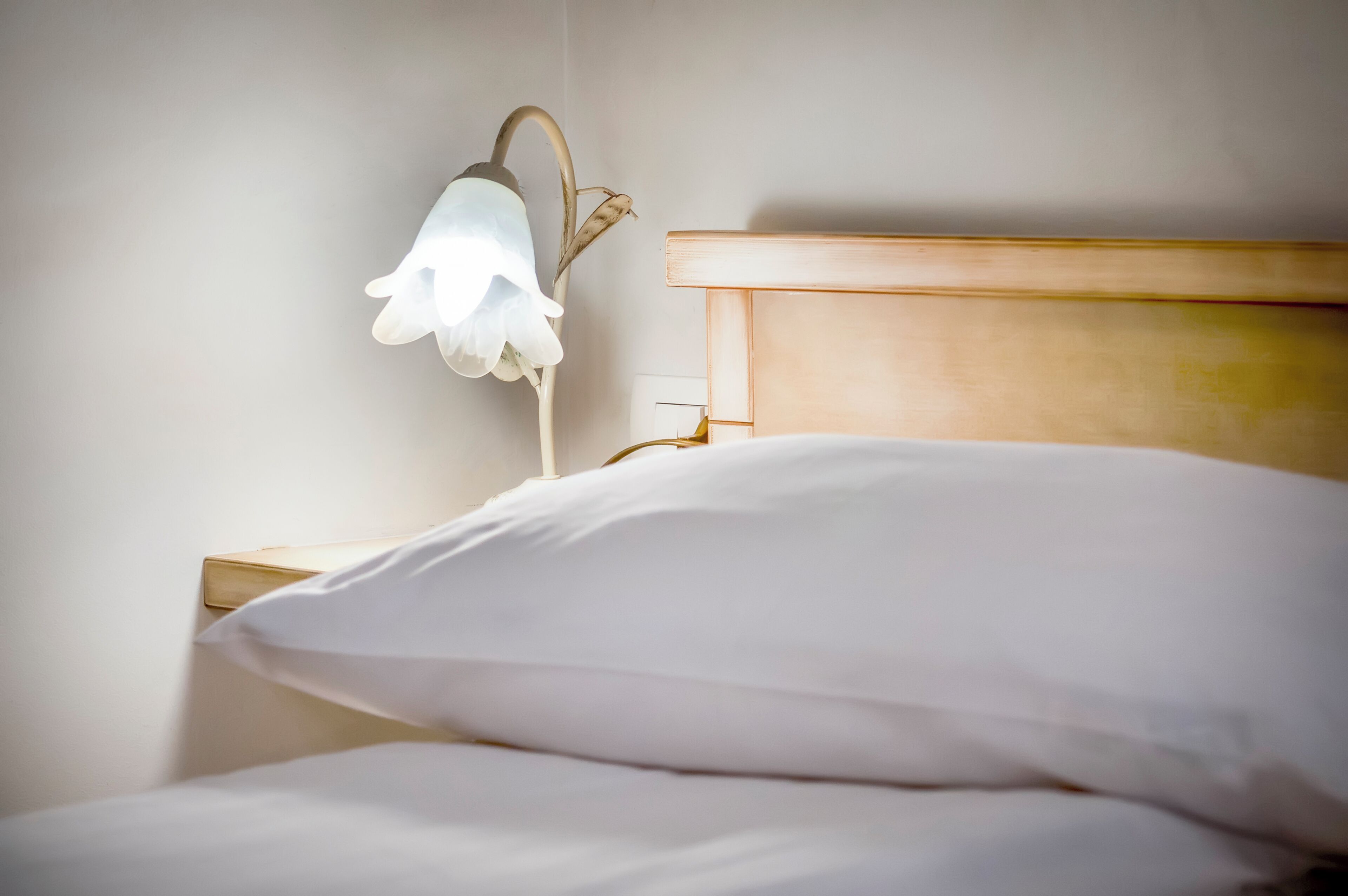 Un lit avec du linge de lit blanc et une tête de lit en bois, agrémenté d’une lampe de chevet avec un abat-jour blanc.