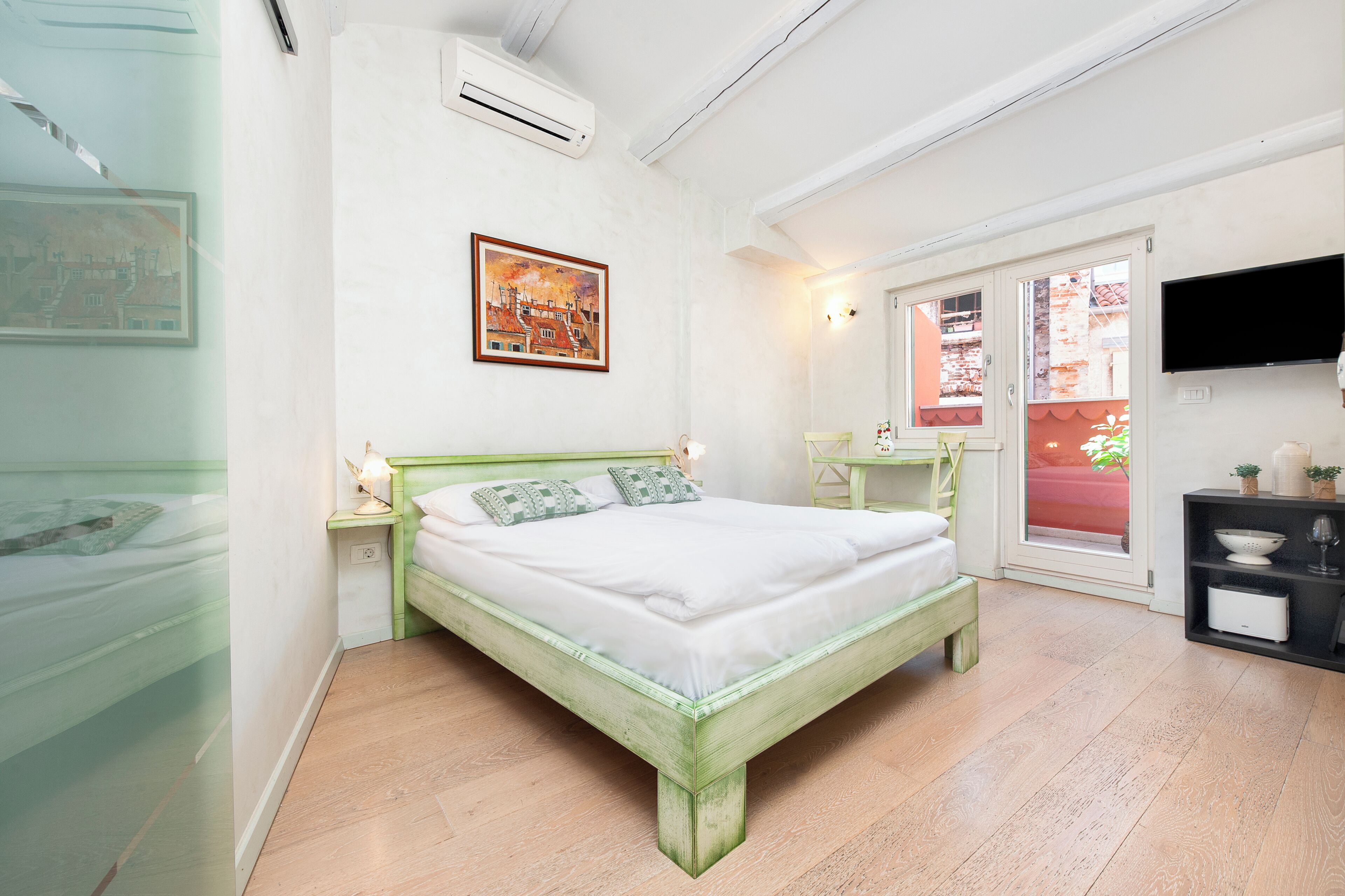 Un dormitorio con cama, mesita de noche, televisión y ventana con vistas a edificios.