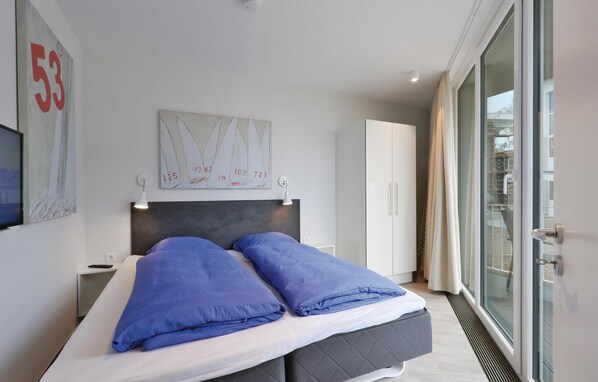 A modern hotel room with a large bed, a headboard with a clock, a television, and a window with a view of buildings. - Dünenvilla 20, Whg 2 (Lübeck Travemünde)