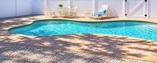 A swimming pool with a brick patio, white outdoor furniture, and a white fence.