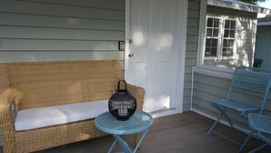 Eine Veranda mit einem Korbsessel, einem kleinen runden Tisch mit einer Laterne und zwei blauen Stühlen.