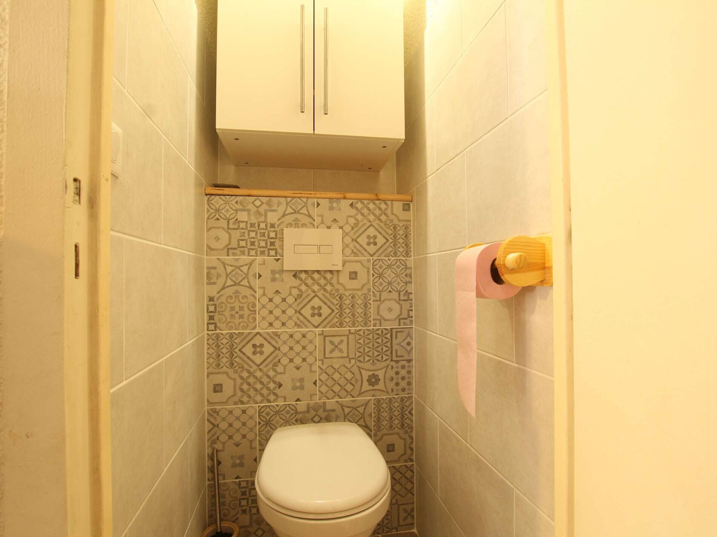 Un baño pequeño con inodoro, baldosas estampadas, un mueble y un dispensador de papel higiénico.