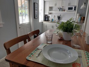 A dining area with a wooden table, chairs, a white plate, a glass, a fork, a knife, a potted plant, and a microwave in the background.
