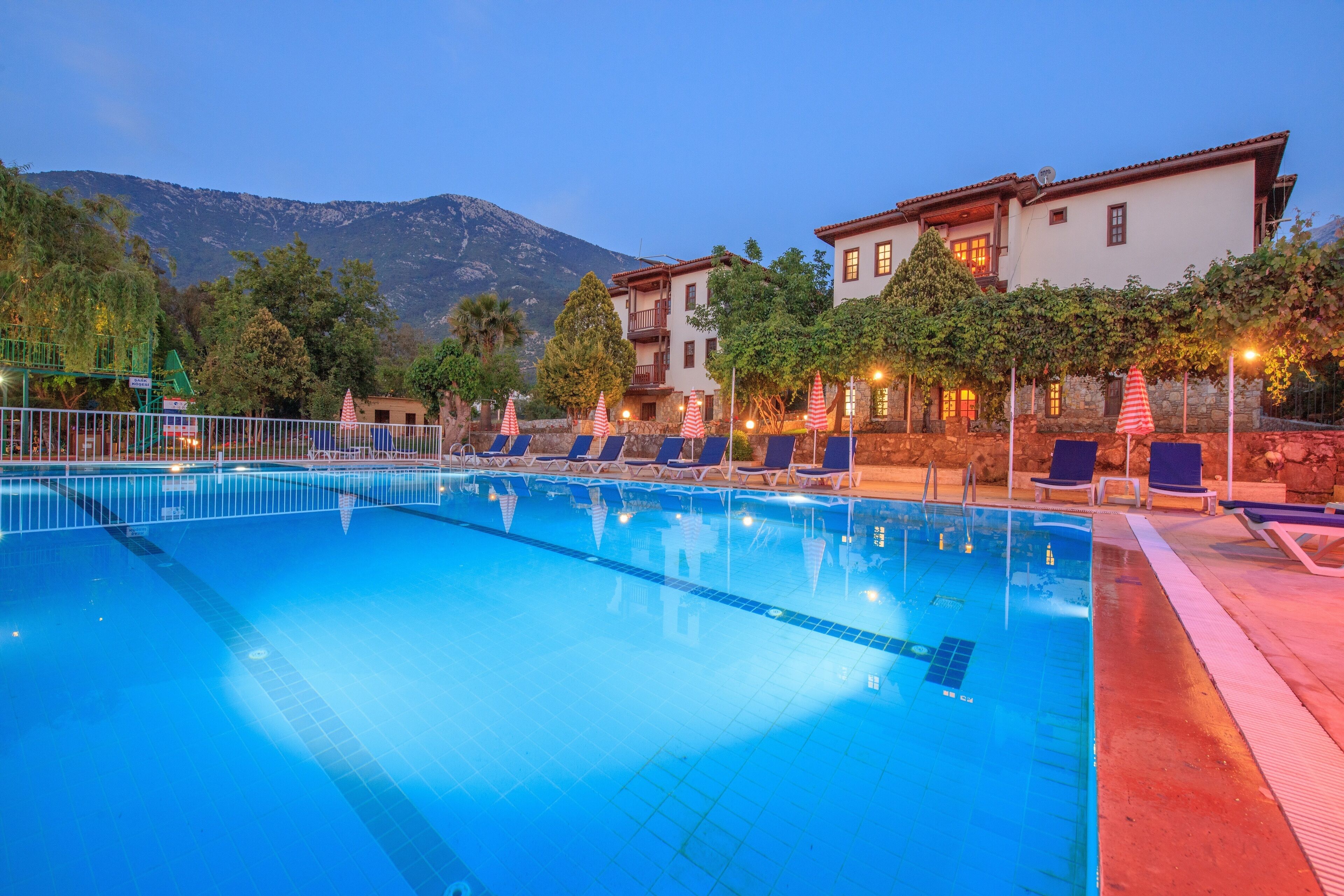 A swimming pool with lounge chairs and umbrellas, surrounded by buildings and trees, with mountains in the background.