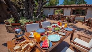 An outdoor dining area with wooden tables and chairs, surrounded by potted plants and a large tree.
