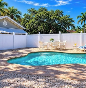 A swimming pool with a brick patio, white outdoor furniture, and a white fence.