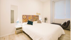 Un dormitorio con una cama, mesitas de noche, un cuadro en la pared, una lámpara de noche, un ventilador de pie y una ventana con persianas.