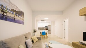Una sala de estar moderna con un sofá, una mesita y un ventilador. Se observa una cocina al fondo, con un refrigerador y un microondas.
