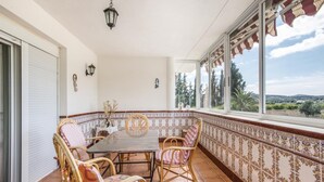 Un patio soleado con una mesa redonda y sillas de mimbre, un muro con azulejos y un diseño decorativo, y vistas a árboles y colinas a través de amplios ventanales.