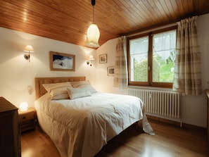 A bedroom with a wooden ceiling, a bed with white bedding, a nightstand, a wall-mounted lamp, and a window with curtains. - Spacious, Bright 3*** Apartment with WIFI (La Clusaz)