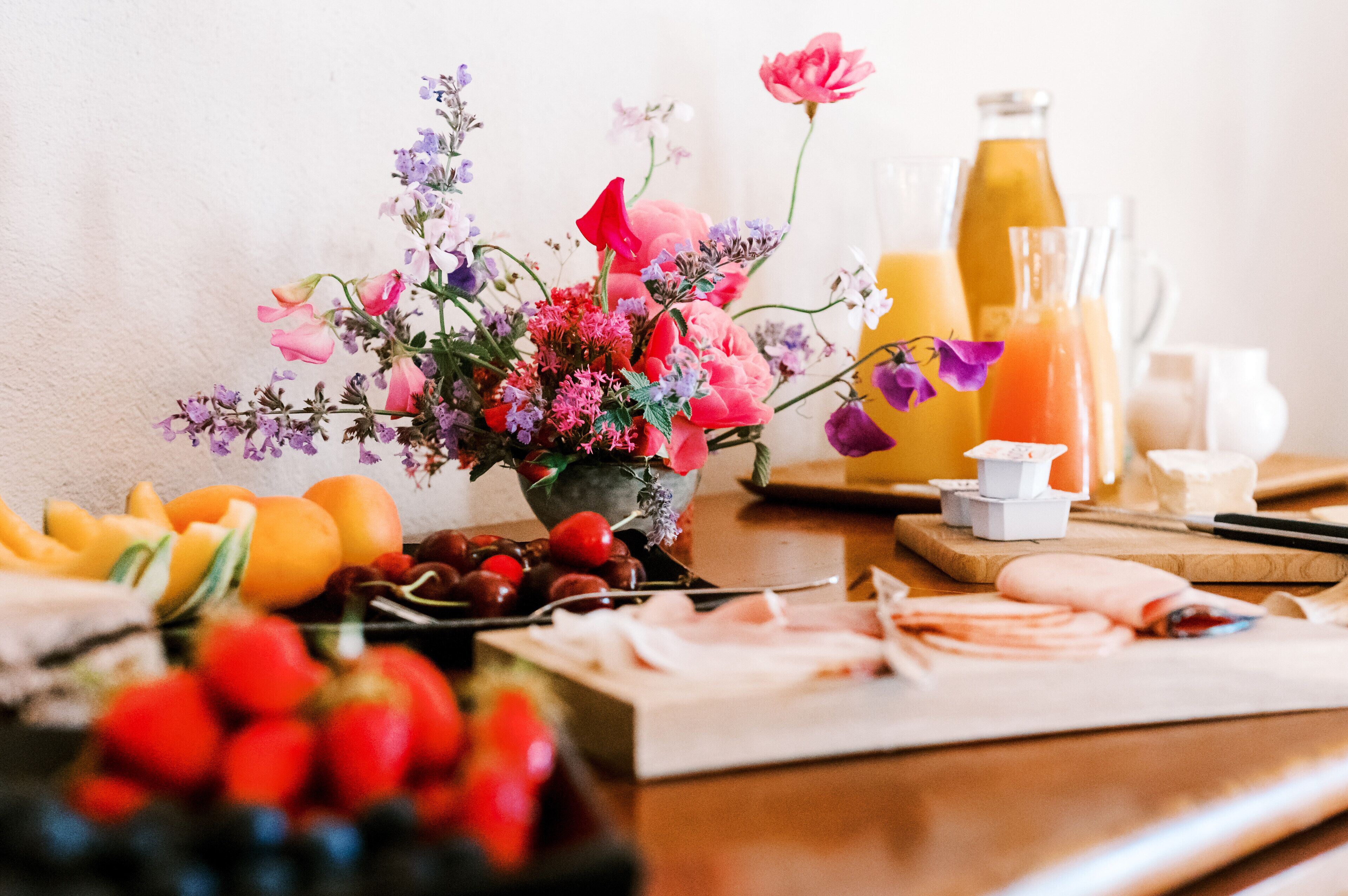 Ett frukostbord med en blomsterdekoration i mitten, färska frukter och en kanna med juice.