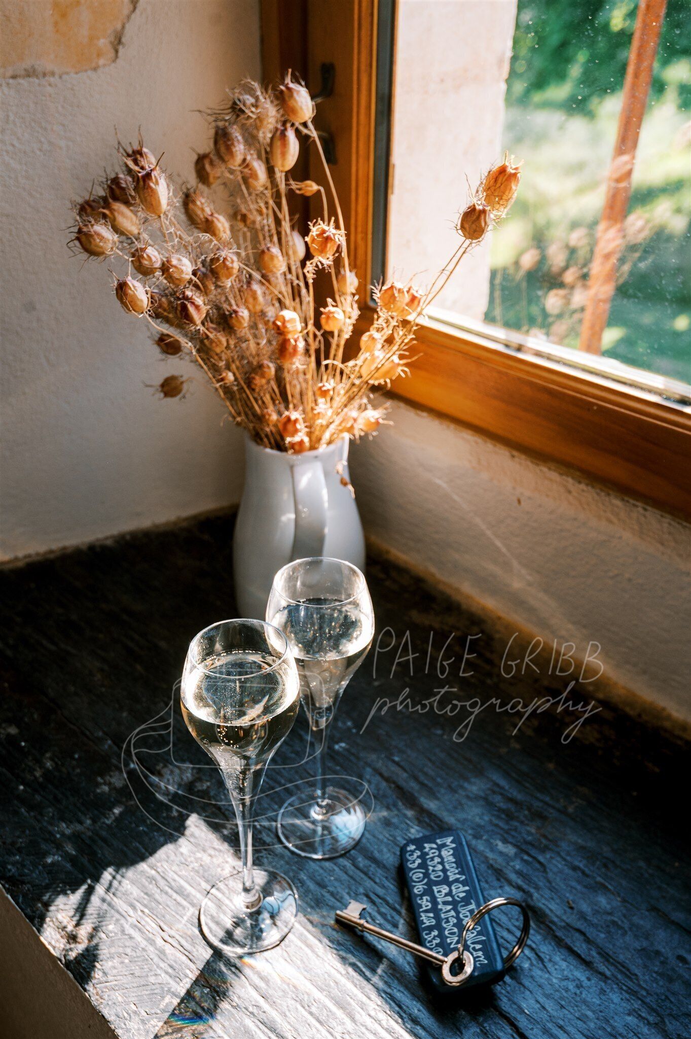 Una ventana con flores secas en un jarrón, dos copas de vino vacías y una llave sobre una superficie de madera.