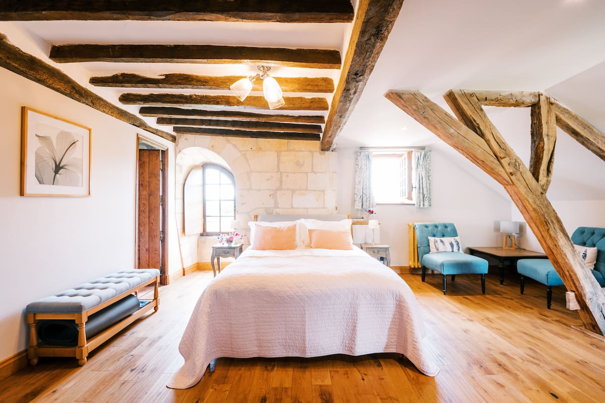 Quarto com uma cama grande, vigas de madeira, um banco e duas poltronas.