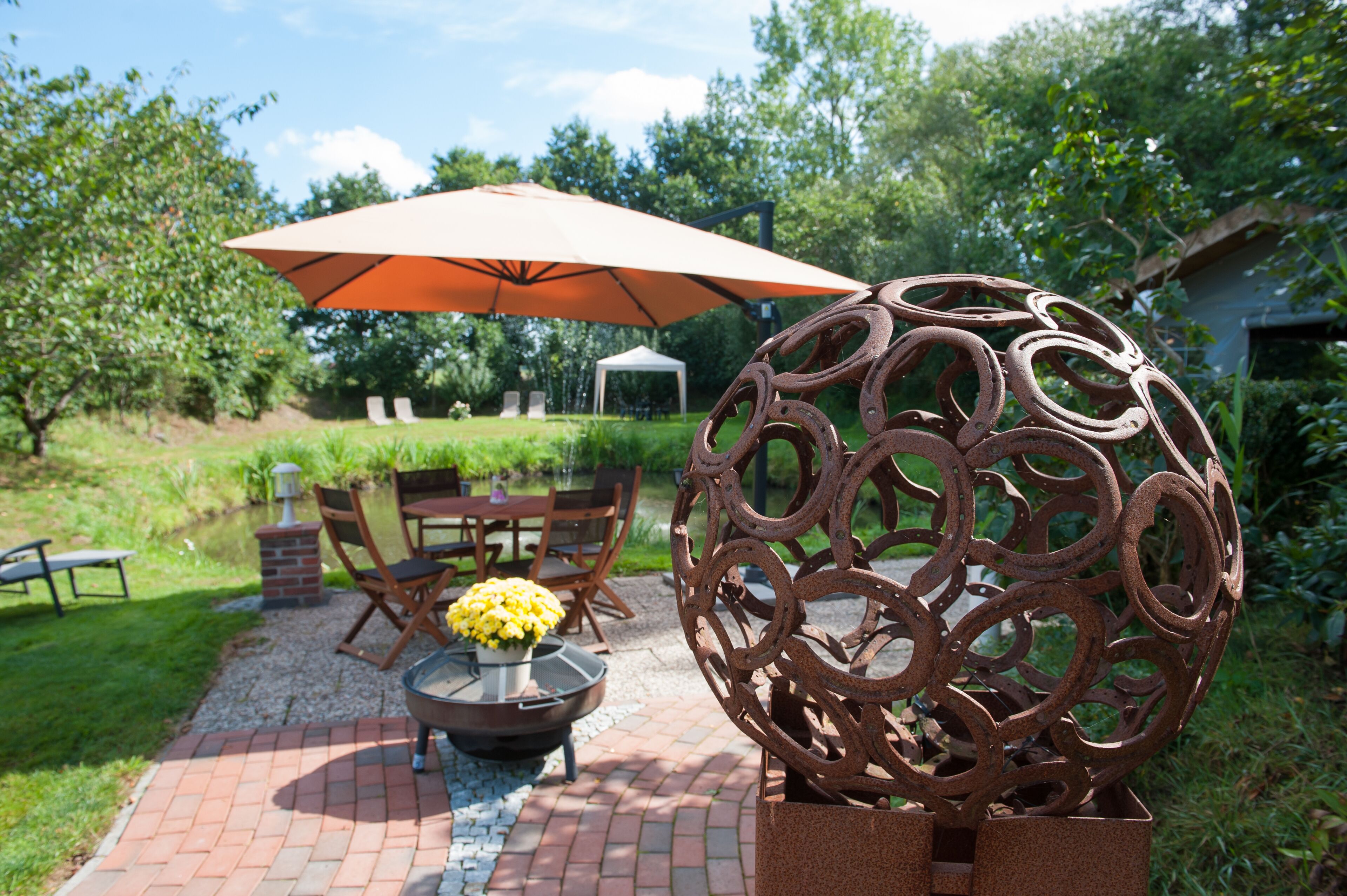 An outdoor patio with a brick pathway, a round metal sculpture, and a table with chairs and an umbrella.