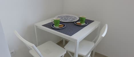 Una pequeña mesa blanca con dos sillas, una taza verde, un plato con una galleta y un mantel individual azul.