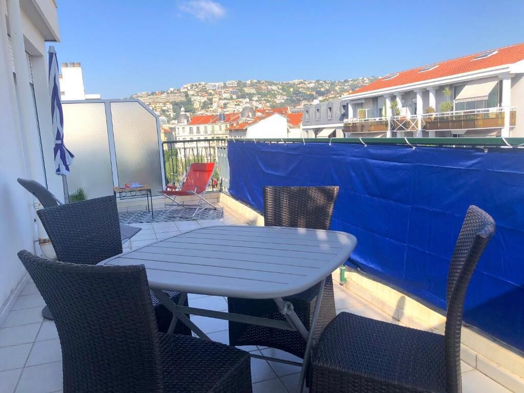 A balcony with wicker chairs and a table, overlooking a cityscape with buildings and a hill.
