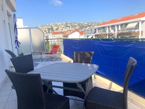 A balcony with wicker chairs and a table, overlooking a cityscape with buildings and a hill.