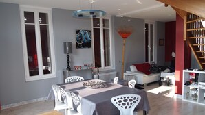 Un coin repas avec une table dressée pour quatre personnes, des chaises blanches, un lustre et, au fond, un canapé rouge et blanc.