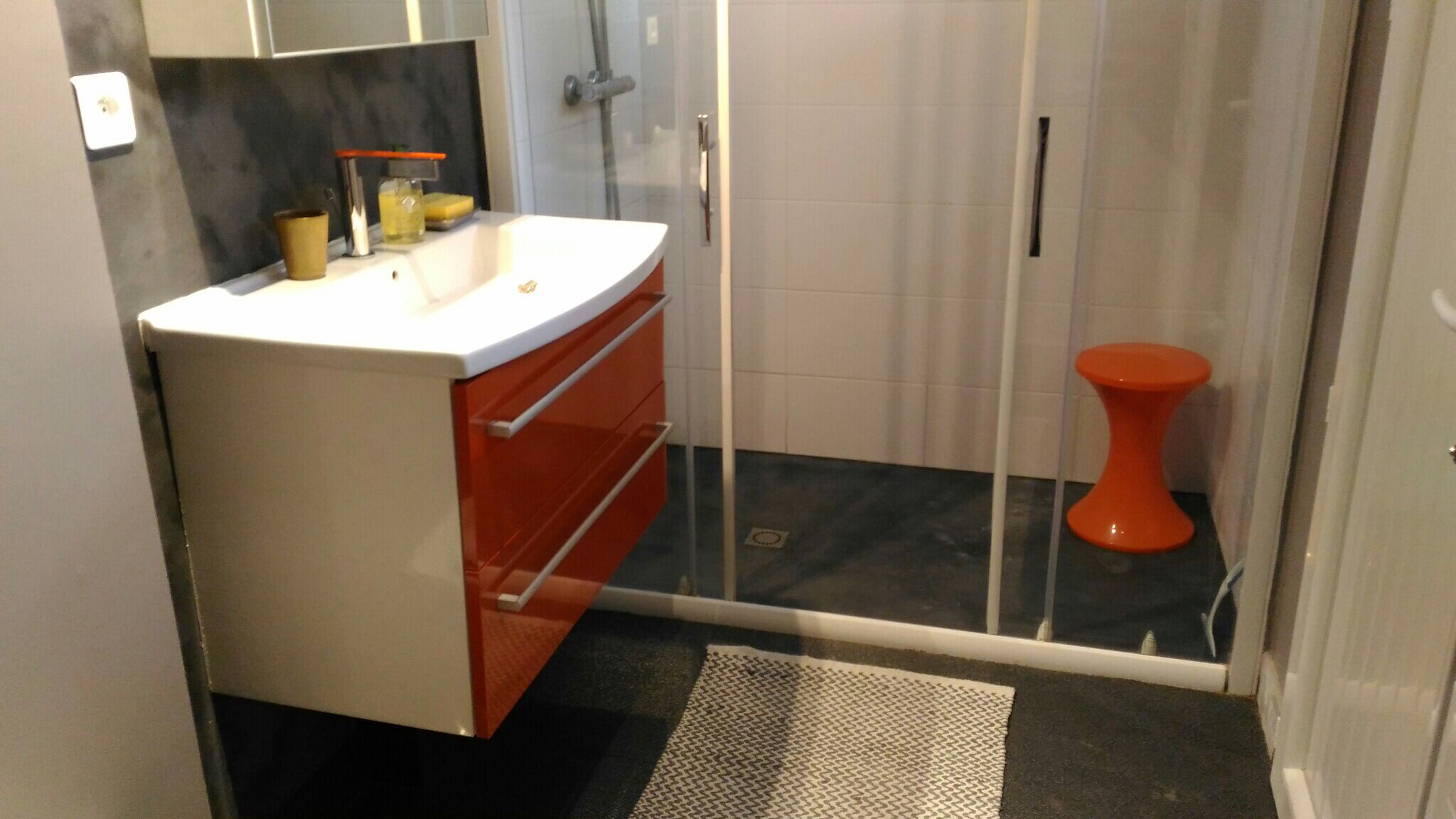Un baño con lavamanos blanco, mueble naranja y mampara de vidrio.