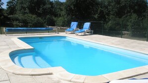Uma piscina retangular com borda curva, rodeada por um deck revestido de azulejos. Há duas espreguiçadeiras com capas azuis e uma cerca ao fundo.