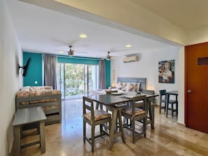 A room with a bed, a dining table with chairs, a ceiling fan, and a large window with curtains.