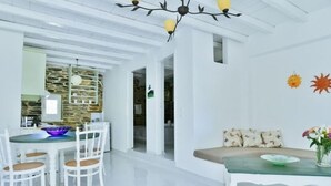 A bright, modern interior with a dining area featuring white chairs and a glass table, a living area with a sofa and cushions, and a kitchen with a stone backsplash. - Cozy 2-bedroom House in Tinos (Tinos)