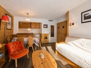 A cozy room with a wooden dining table, a red chair, a wooden coffee table, and a bed with a white blanket. - 2** Apartment for 6, South, near slopes and shops (La Clusaz)
