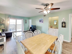 A spacious living area with a wooden dining table, white chairs, a ceiling fan, and a view through a sliding glass door to an outdoor balcony.