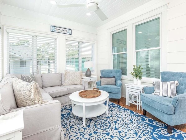 A bright, sunlit room with a white ceiling fan, a beige sofa, a blue armchair, and a white coffee table.