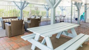 Een terras buiten met een witte picknicktafel, rieten meubels en een overdekt gedeelte met een witte schutting.