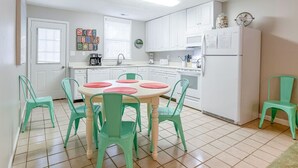 A compact kitchen with white cabinets, a central island with pink and green chairs, a refrigerator, and a coffee maker. - Latitude Adjustment | Pet Friendly, Prime Location (Tybee Island)