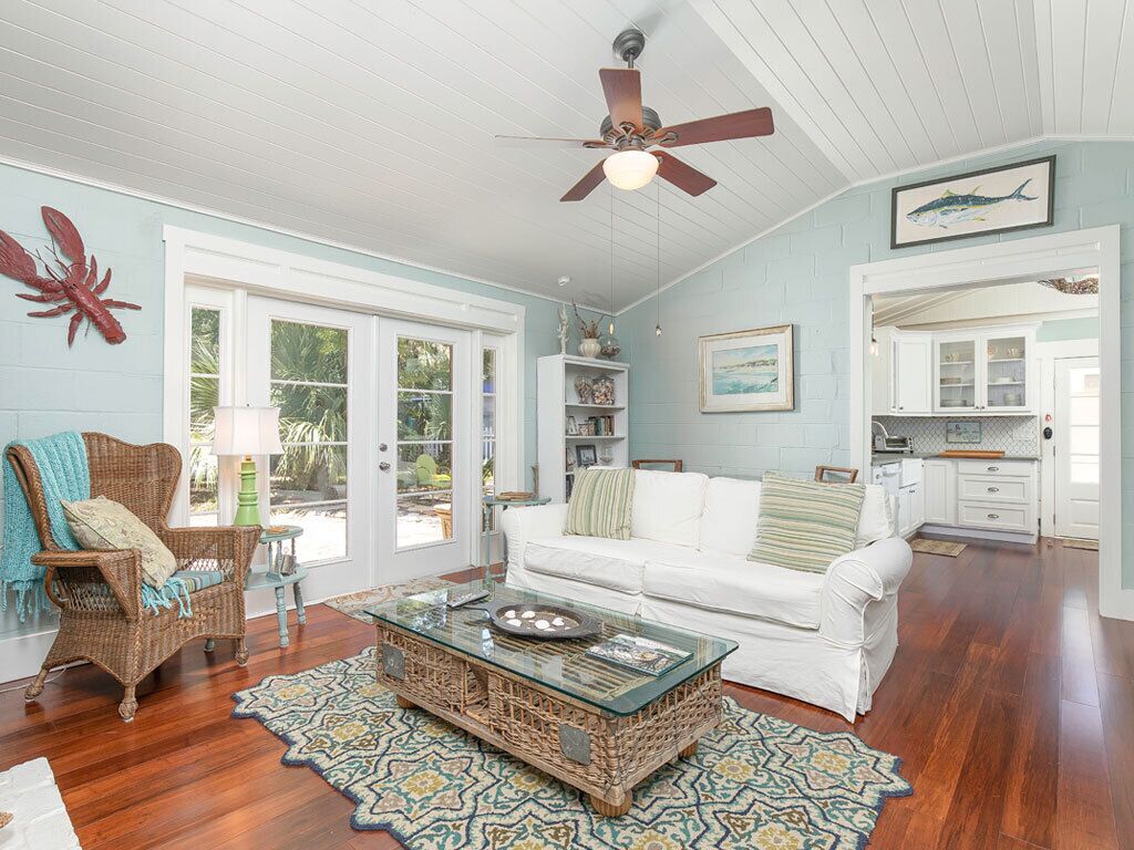 A spacious living room with a white sofa, wicker armchair, and a glass coffee table. There is a ceiling fan, a large window, and a kitchen area visible through an open door.