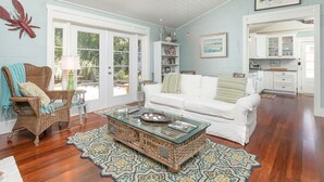 A spacious living room with a white sofa, wicker armchair, and a glass coffee table. There is a ceiling fan, a large window, and a kitchen area visible through an open door. - Sunny Daze | Large Patio + Fire Pit! (Tybee Island)