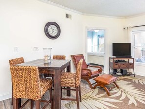 A dining area with a wooden table and woven chairs, a television, and a decorative clock on the wall. - Sandpiper 107 | Ocean Views, 20 Yards to Beach! (Tybee Island)